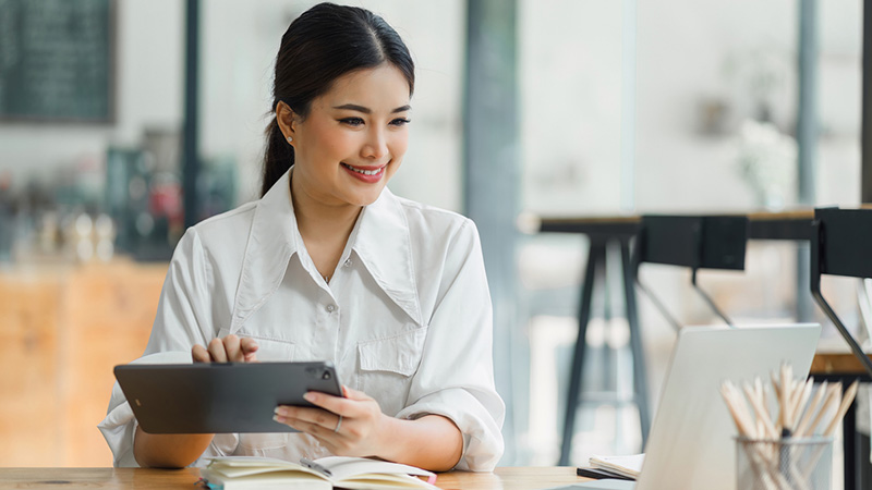 woman sitting at a desk holding a tablet looking at a laptop